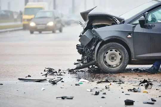 Front-end damage of a black car after a highway collision, with debris scattered on the road.