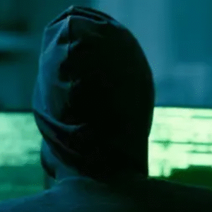 A person in a dark hoodie facing multiple computer screens with green code, symbolizing hacking or cybercrime in a dimly lit room.