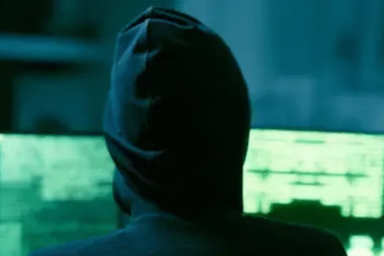 A person in a dark hoodie facing multiple computer screens with green code, symbolizing hacking or cybercrime in a dimly lit room.
