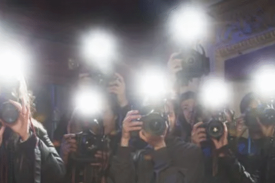 A group of photographers taking pictures with camera flashes on a red carpet event, standing behind a velvet rope barrier.
