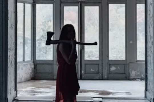 Silhouetted woman in a red dress holding an axe inside an abandoned, dimly lit building with tall glass doors in the background.