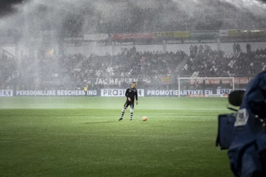 Football player on a floodlit field during a live match as sprinklers spray water; cameramen and spectators in the background.