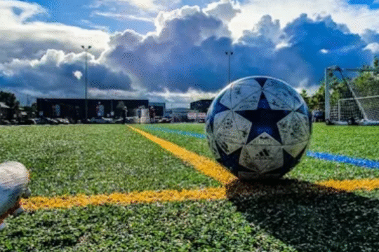 A football and cleats on a green turf field under a dramatic cloudy sky with goalposts in the background.