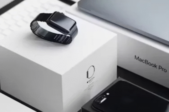 A collection of Apple products including a MacBook Pro, Apple Watch, iPhone, and keyboard placed aesthetically on a white desk.