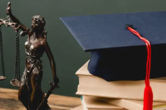 Lady Justice statue beside a stack of law books topped with a graduation cap on a wooden table.
