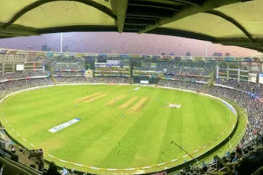 Panoramic view of a packed cricket stadium during a live match, with bright lights, lush green field, and cheering crowd under a twilight sky.