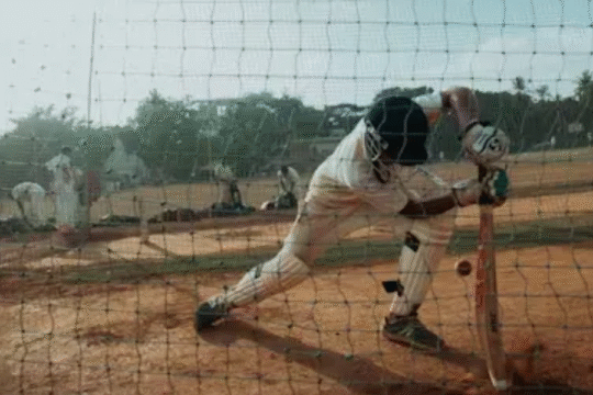 Young cricketer practicing a defensive shot inside a training net on a dusty outdoor field, surrounded by teammates and coaches.