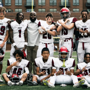 A high school American football team poses together on the field after a game, wearing white and maroon "PREP" uniforms. Players and coaches smile and make celebratory gestures, with a scoreboard in the background showing the final score.
