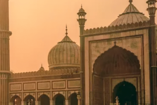 A majestic mosque at sunset with intricate Mughal architecture, featuring domes and a tall minaret, set against a warm orange sky.