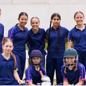 A girls' school cricket team posing for a group photo in blue uniforms, with some players wearing helmets and holding cricket gear, standing in front of a white picket fence on a grassy field.