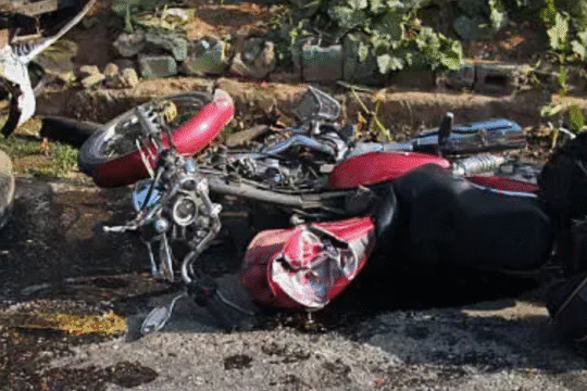 A damaged red motorcycle lies on the road next to an overturned vehicle, surrounded by debris and oil stains, indicating a serious traffic accident.