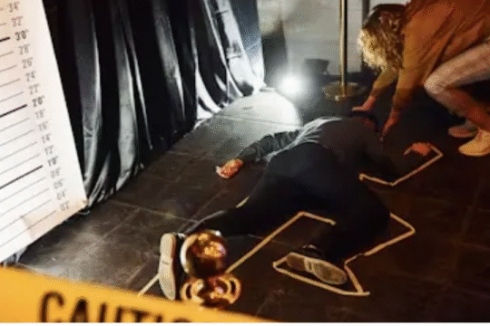 A staged crime scene with a person lying face down inside a chalk outline, while another individual kneels beside them. The area is surrounded by caution tape and dramatic lighting, with a height measurement backdrop visible.