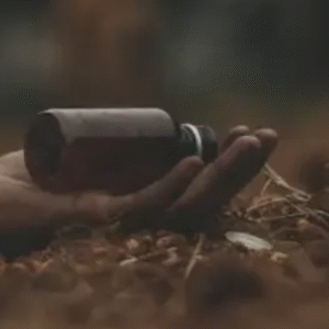 A close-up of a person's hand lying lifeless on the ground, holding a small brown medicine bottle, surrounded by dry leaves and soil, implying a possible overdose or poisoning.
