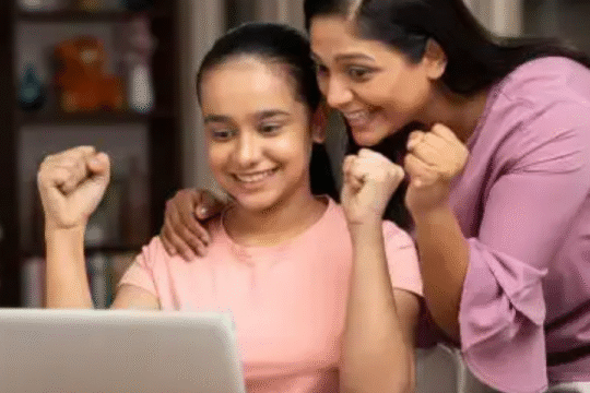 A happy teenage girl celebrating exam results on a laptop with her smiling mother beside her, showing excitement and pride at home.