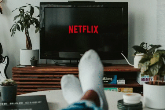 Person relaxing at home while watching Netflix on a TV screen, surrounded by indoor plants and a guitar in the background.