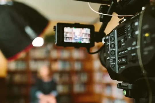 A video camera records a person sitting in front of a bookshelf, with lights.