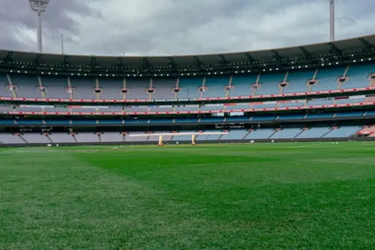 A wide view of an empty cricket stadium with green grass and towering light posts against a cloudy sky.