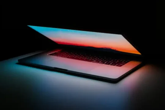 An open laptop in a dark room, screen displaying a colorful sunset.