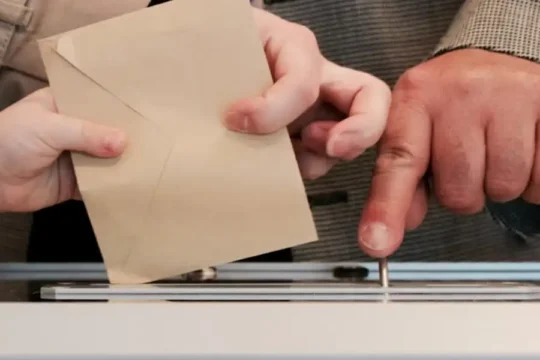 One hand holds a ballot envelope while another hand points to the slot of a ballot box.