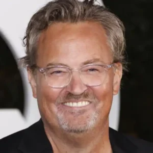 Actor Matthew Perry smiling warmly, wearing glasses and a black jacket.