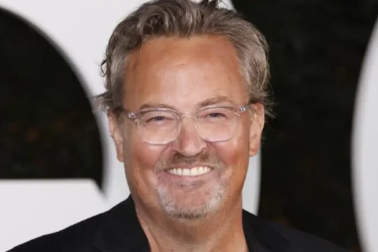 Actor Matthew Perry smiling warmly, wearing glasses and a black jacket.