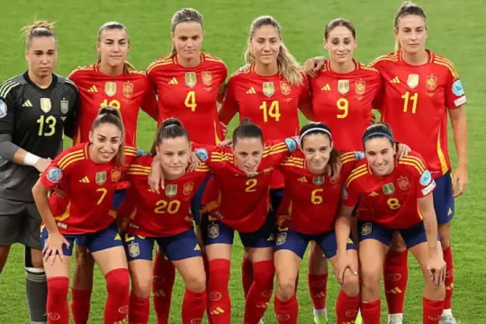 Spanish women's football team posing together in red jerseys on a field.