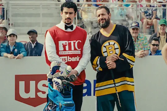 Adam Sandler in a hockey jersey with a caddy on a golf course.