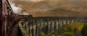 A steam train crossing a large arched viaduct in a misty, mountainous landscape.
