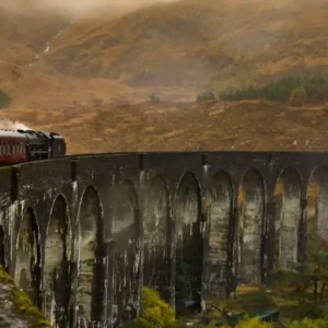 A steam train crossing a large arched viaduct in a misty, mountainous landscape.