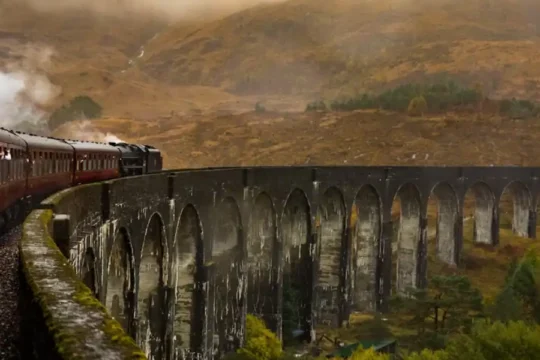 A steam train crossing a large arched viaduct in a misty, mountainous landscape.