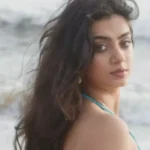 A young woman with long, dark, wavy hair looks over her shoulder at a beach.