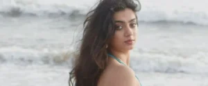 A young woman with long, dark, wavy hair looks over her shoulder at a beach.