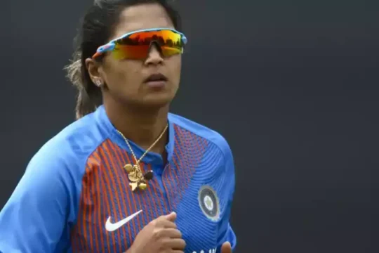 An Indian female cricketer in blue jersey and sunglasses, looking focused.