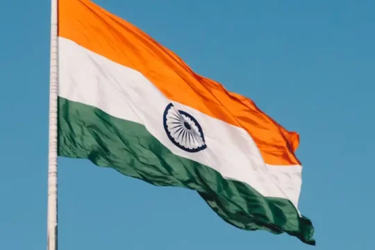 A large Indian flag (Tiranga) flapping in the wind against a clear blue sky.
