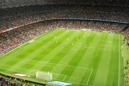A wide, high-angle view of a brightly lit football stadium filled with spectators during a night match.