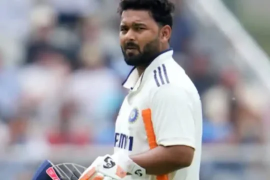 Cricketer Rishabh Pant in white uniform, holding his helmet and bat on the field.