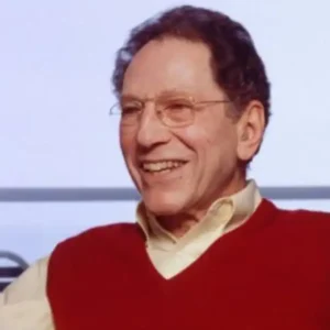 An older man with glasses in a red sweater vest, smiling and gesturing with his hands.