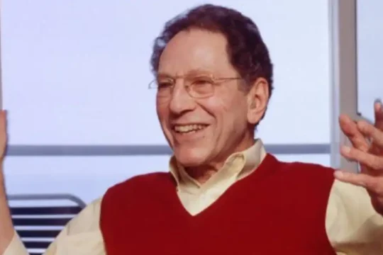 An older man with glasses in a red sweater vest, smiling and gesturing with his hands.