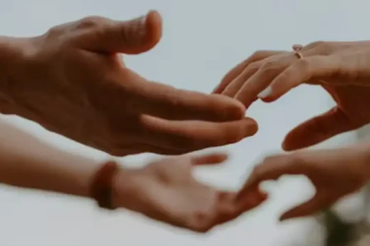 Four hands reaching towards each other, with one wearing a ring, against a bright sky.