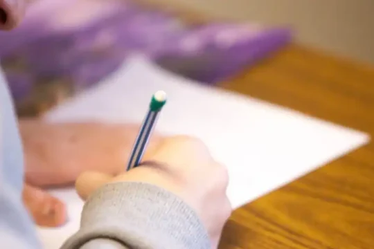 A person's arm and hand holding a pen and writing on a white paper on a wooden desk
