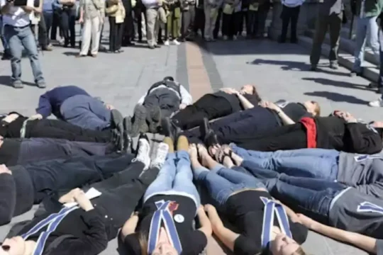 People lying on the ground in a circle, performing a "die-in" protest, surrounded by onlookers and media