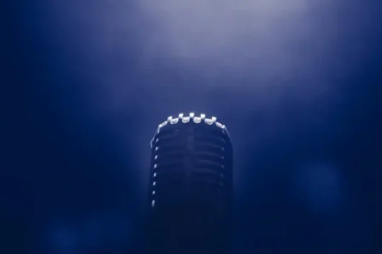 A lone vintage microphone on a stage, illuminated by a spotlight from above against a dark blue background.