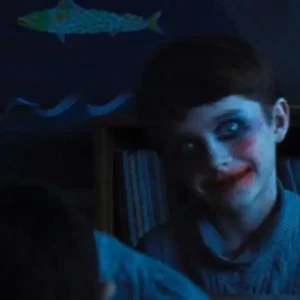 A pale-faced boy with red lips and dark eyes, looking menacingly over another child's shoulder in a classroom.