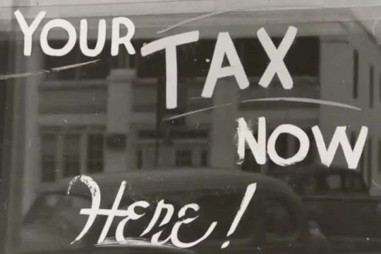 A monochrome image showing a window with "PAY YOUR TAX NOW Here!" painted on it