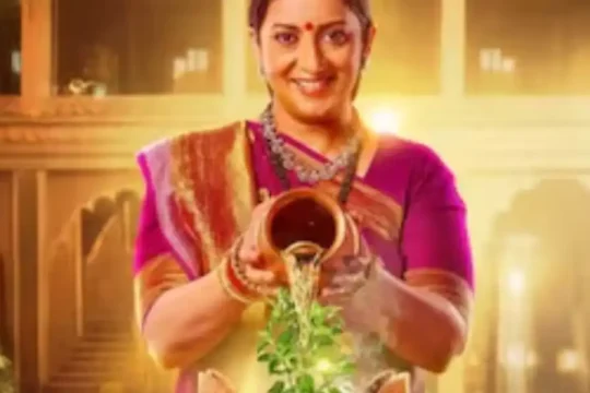 A smiling Indian woman in traditional attire pours water from a pot onto a tulsi plant.