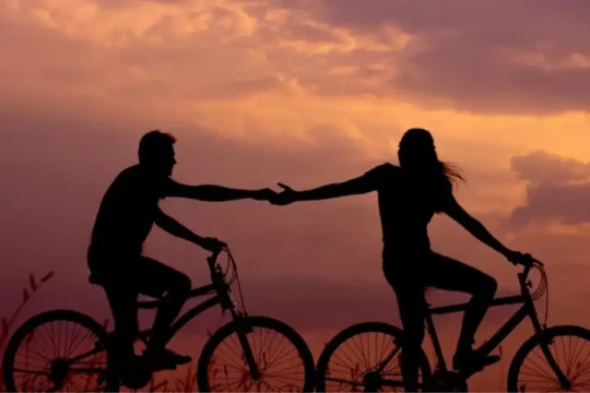 Silhouettes of a couple on bicycles holding hands against a vibrant sunset sky.