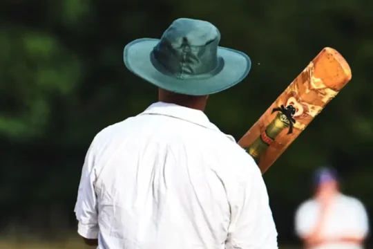 A cricketer from behind, wearing a white shirt and a green hat, holding a bat, with another player blurred in the background