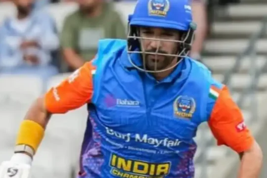 A cricketer in a blue and orange India Champions jersey, helmet, and batting pads, holding a bat and running.