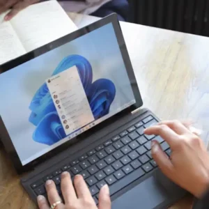 Two people are at a wooden table, one typing on a laptop running Windows 11.