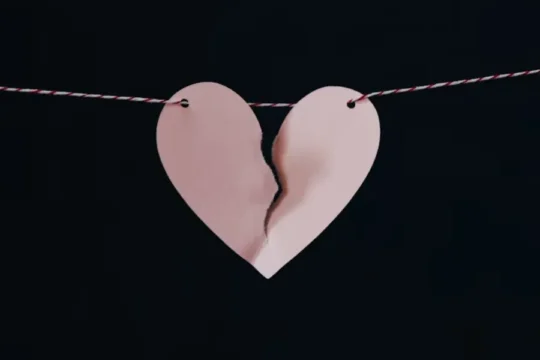A pink paper heart, torn in half, hangs on a string against a dark background.